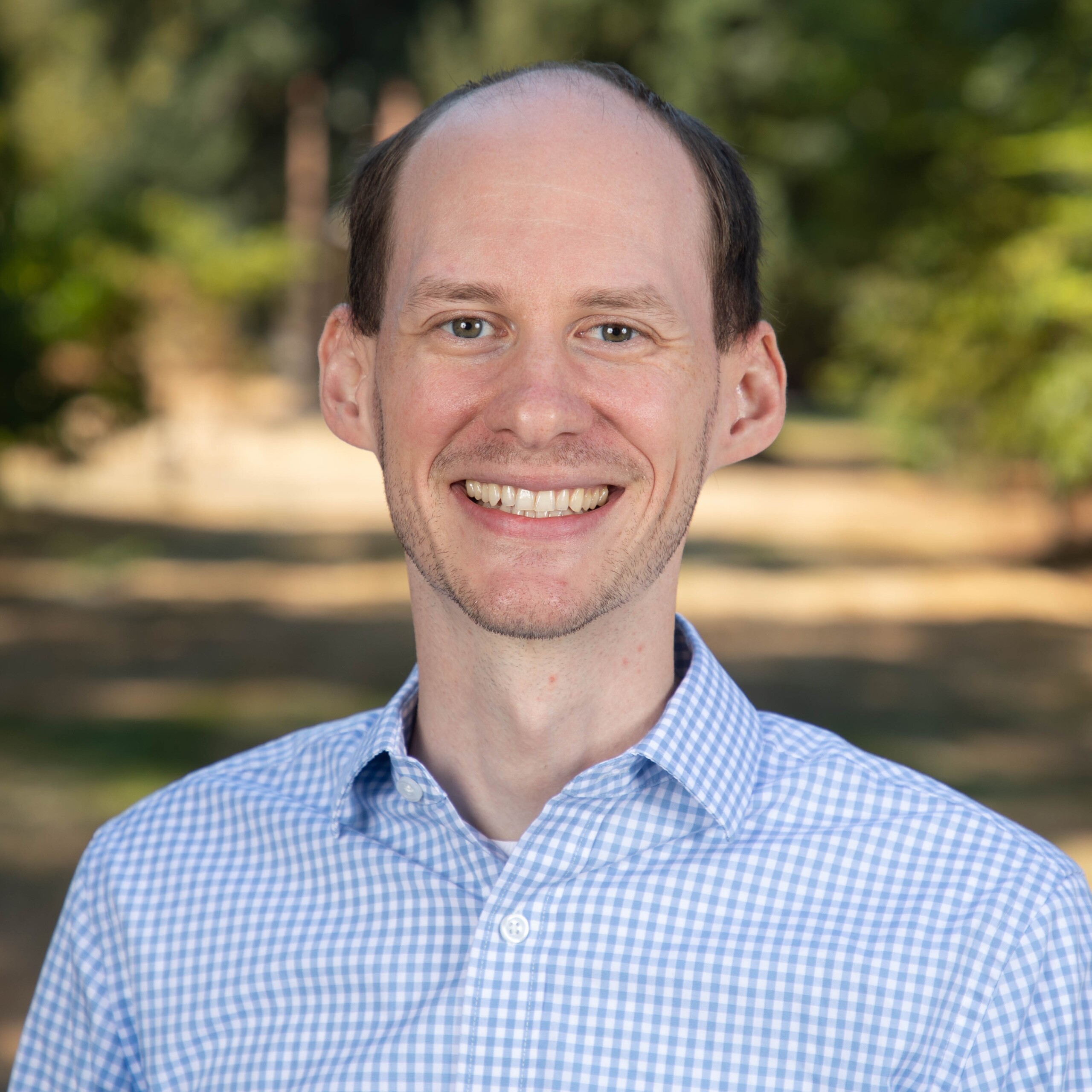 Micah Miller smiles in a checkered blue and white dress shirt with blurry trees in the background.