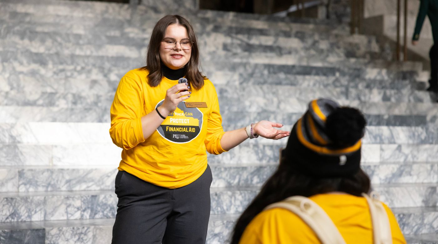 A PLU student in a yellow shirt that reads, "My voice can protect financial aid" holds a small microphone while standing on the steps of the Washington State Capitol Building.