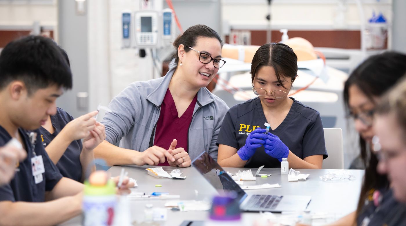 Clinical Instructor of Nursing Kristina Kenning, left in maroon shirt and glasses, leads a nursing student holding a needle during a course titled "Clinical Practicum II Lab."
