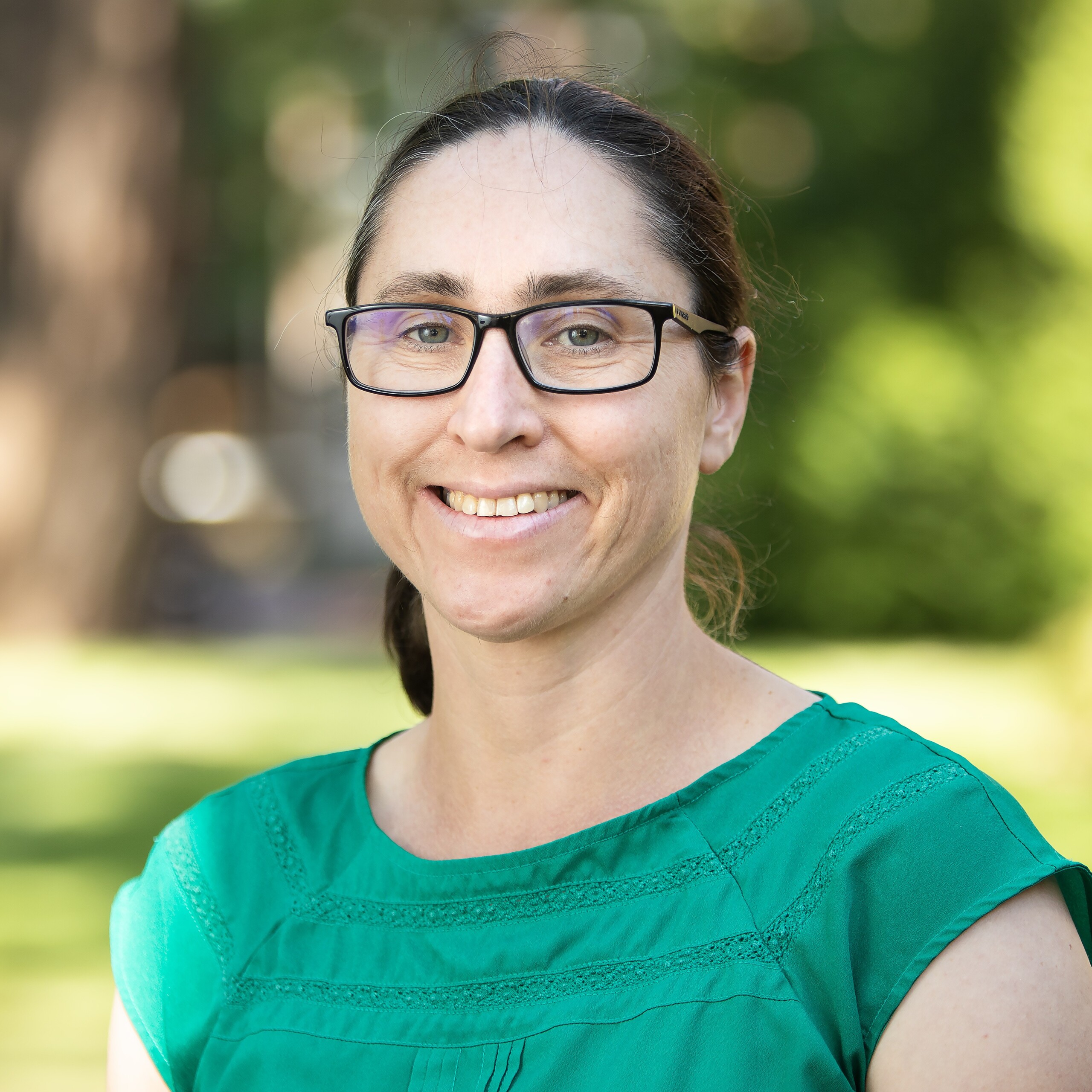 Sara Finley smiles while wearing a green shirt and glasses. Green trees and grass are blurry in the background.
