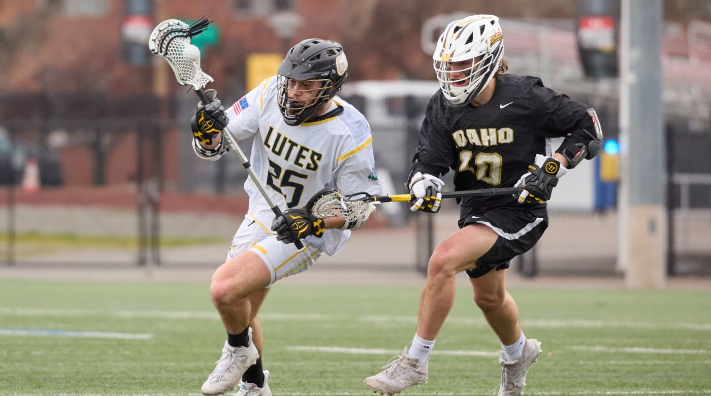 A Idaho Lacrosse player in a black jersey and white helmet chases closely behind a PLU Lacrosse player with the ball in a white jersey and gray helmet.