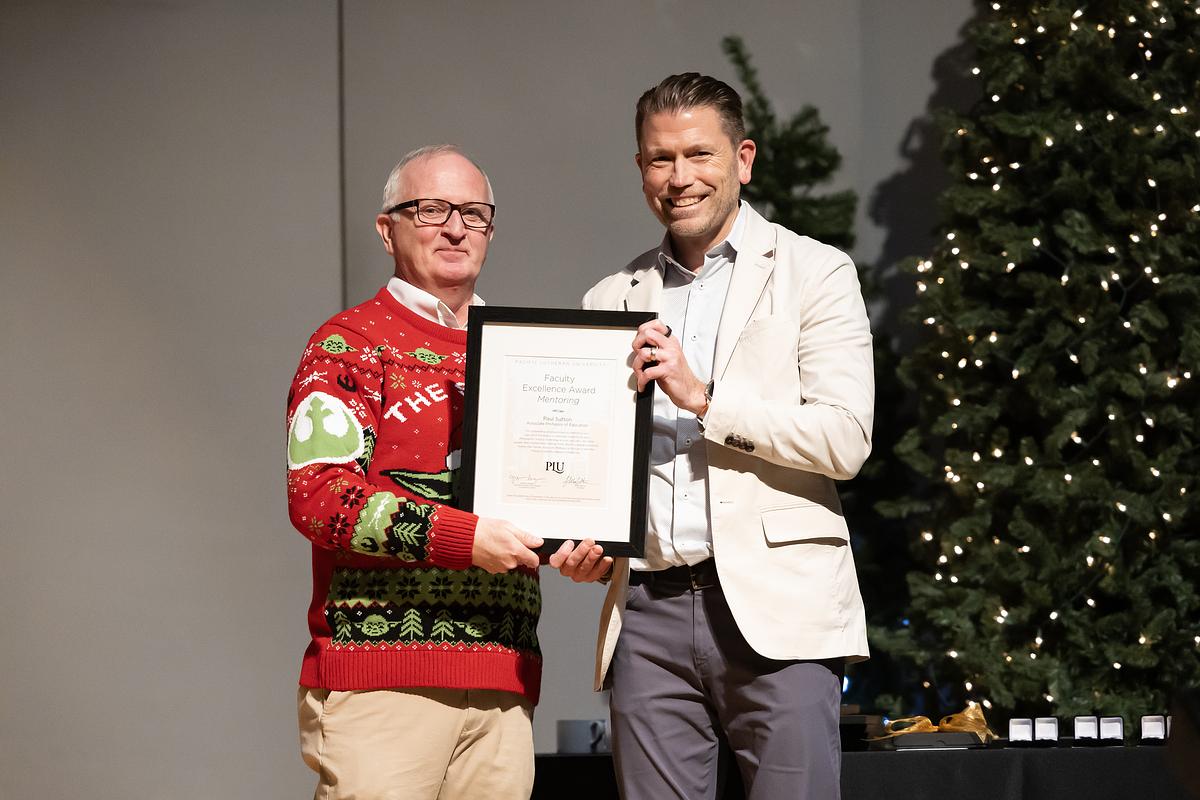 PLU President Allan Belton, in a red holiday sweater, awards professor Paul Sutton, white blazer, with an award during the 2024 Staff and Faculty Celebration.