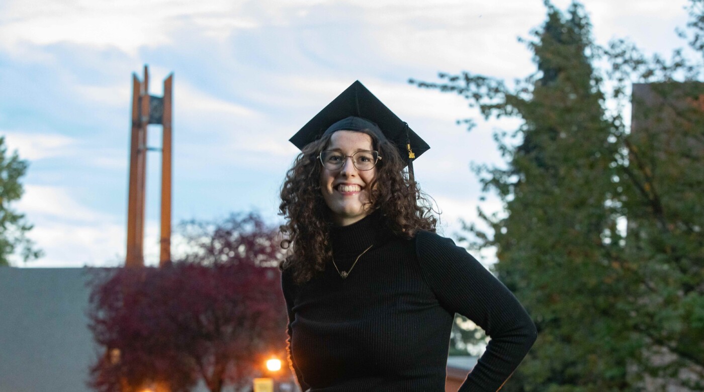 Student Ava Foley wears a graduation cap and smiles at the camera outside of a building on PLU's campus.