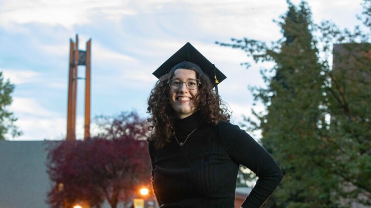 Student Ava Foley wears a graduation cap and smiles at the camera outside of a building on PLU's campus.