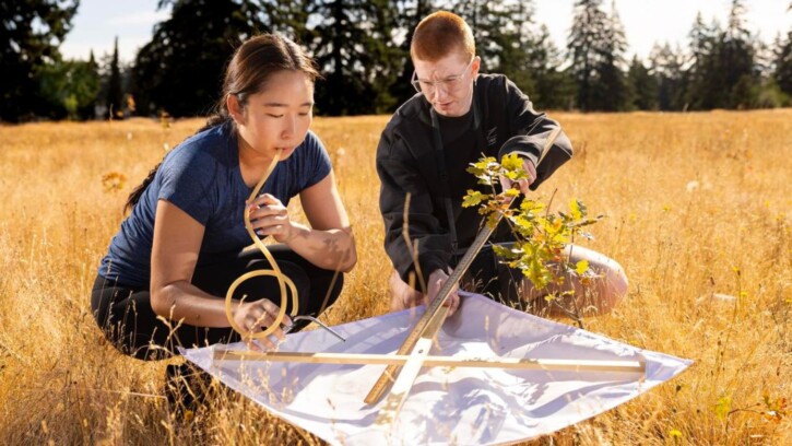 Two students sit in a field looking over a white tarp. The student in the foreground has a rubber tube in her mouth and the student in the background is closely examining the surface of the white tarp.