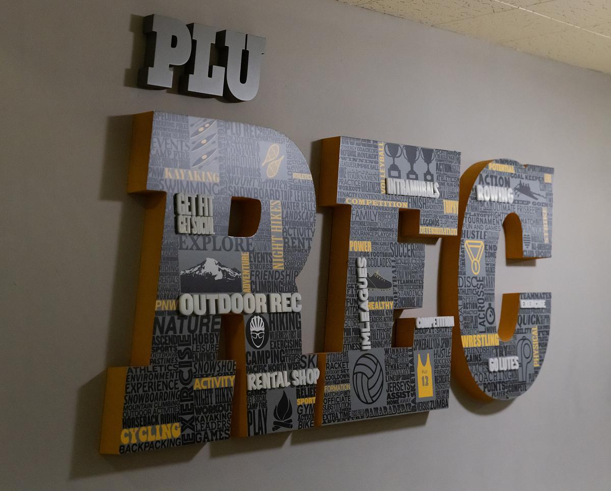 Collage of outdoor recreation and activity themed text and graphics fill a sign that reads, "PLU Rec" in large letters hanging on a wall.