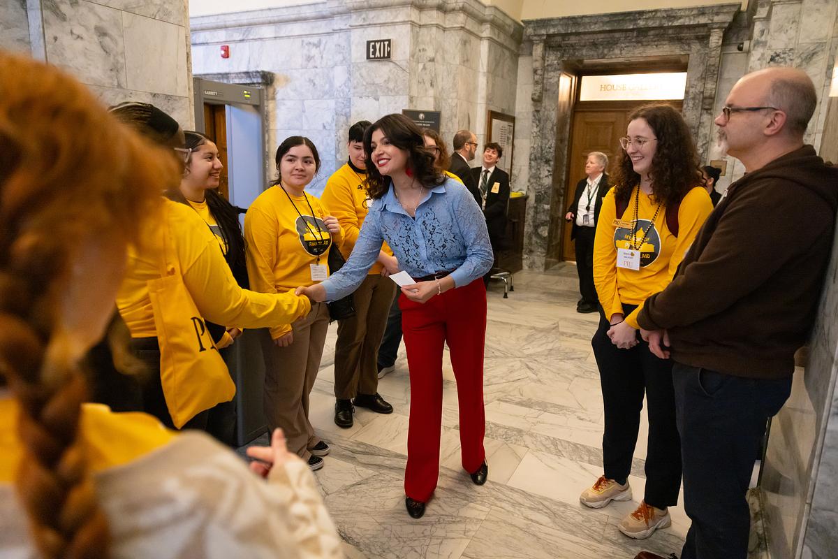 Washington State Representative, Sharlett Mena, in red pants and a blue top, shakes hands and chats with PLU students in yellow shirts.