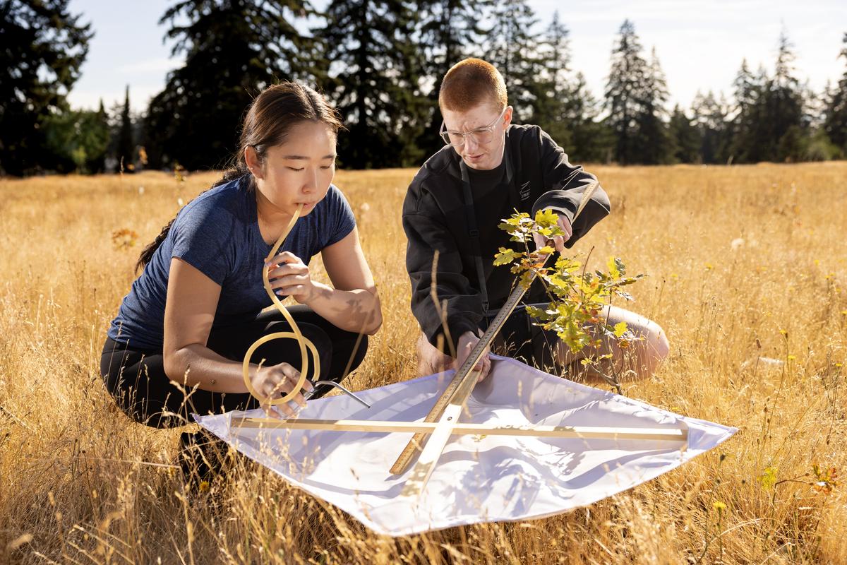 Two students sit in a field looking over a white tarp. The student in the foreground has a rubber tube in her mouth and the student in the background is closely examining the surface of the white tarp.