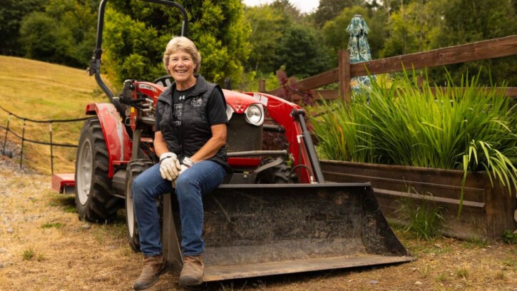 Assistant Softball Coach Cindy Van Hulle, wearing blue jeans, brown boots, gloves, and a black sweater, smiles as she sits on her tractor at her home/farm on Friday, July 17, 2026, at PLU. (PLU Photo / Sy Bean)