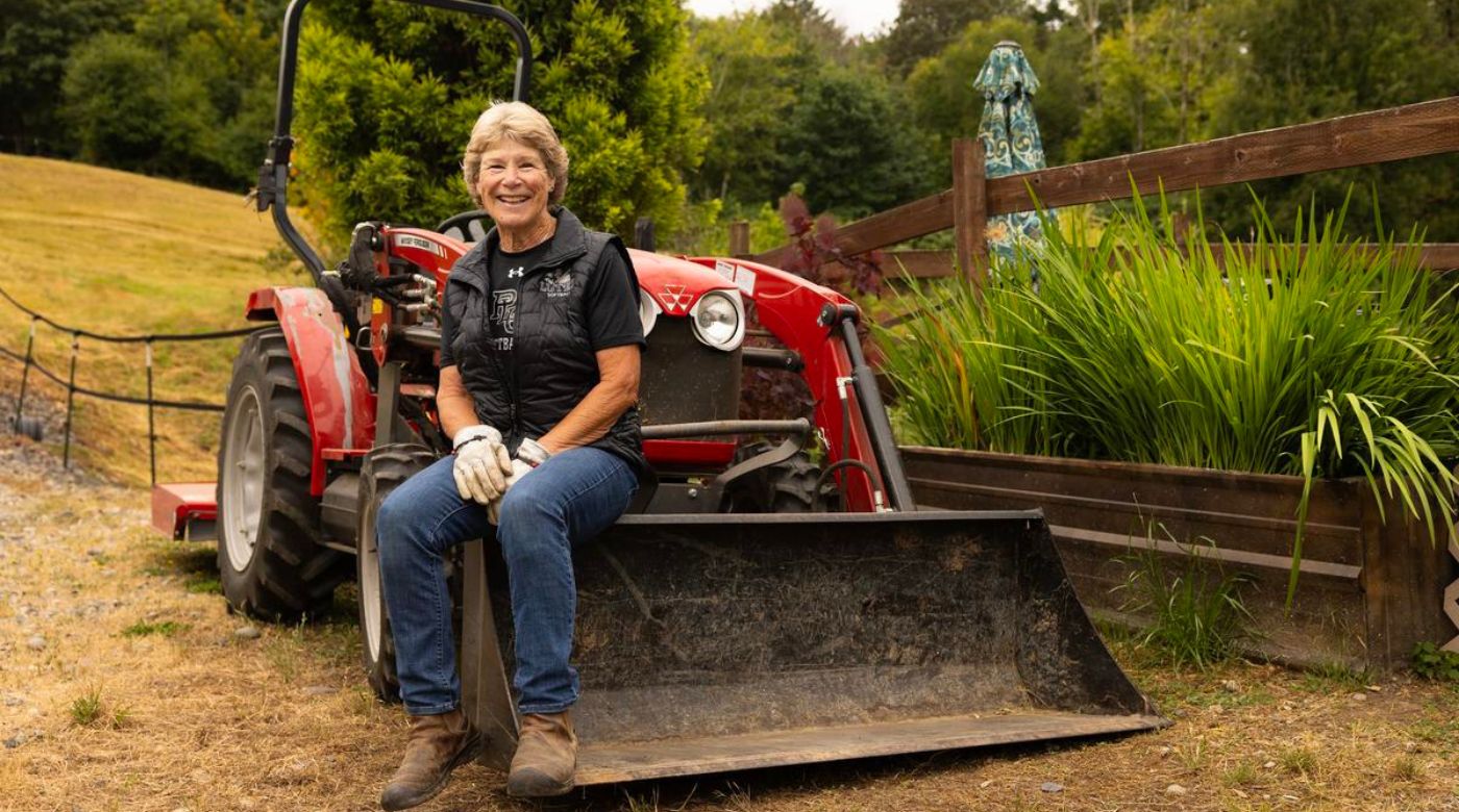 Assistant Softball Coach Cindy Van Hulle, wearing blue jeans, brown boots, gloves, and a black sweater, smiles as she sits on her tractor at her home/farm on Friday, July 17, 2026, at PLU. (PLU Photo / Sy Bean)