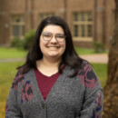 Person with long hair wearing glasses and cardigan smiling outdoors on campus