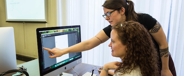 Person standing and pointing something out on a computer screen for a seated person