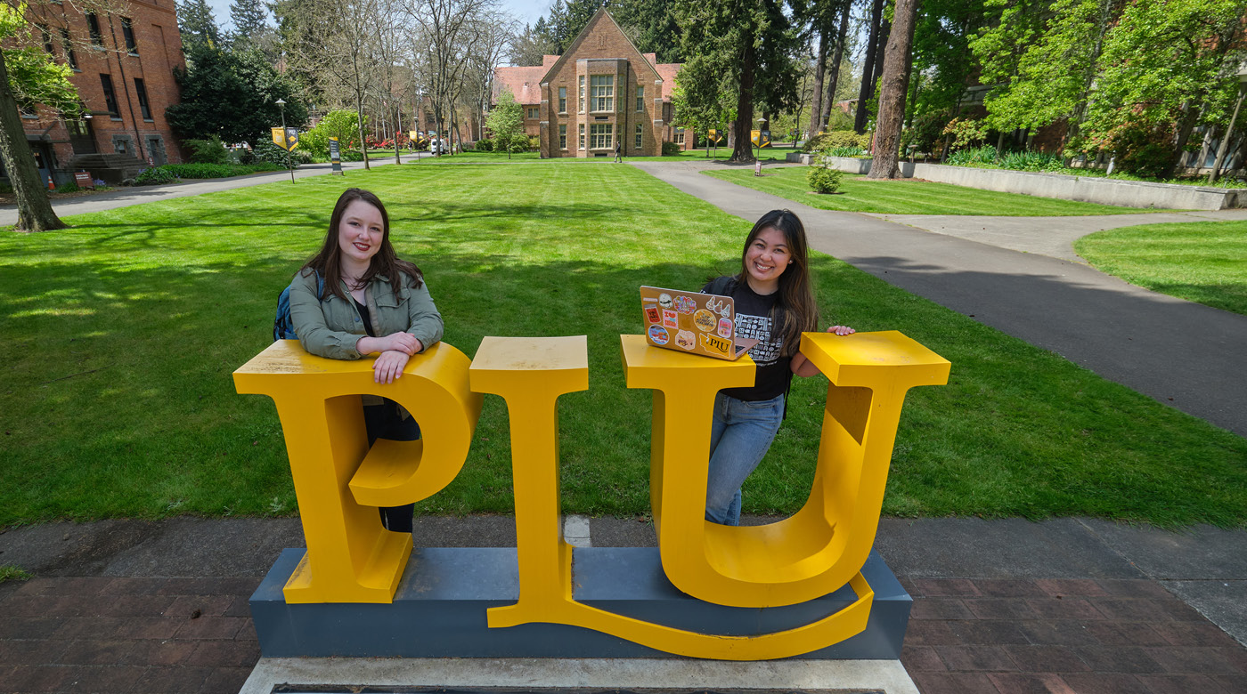 Two students standing smiling in front of the PLU sign on upper campus