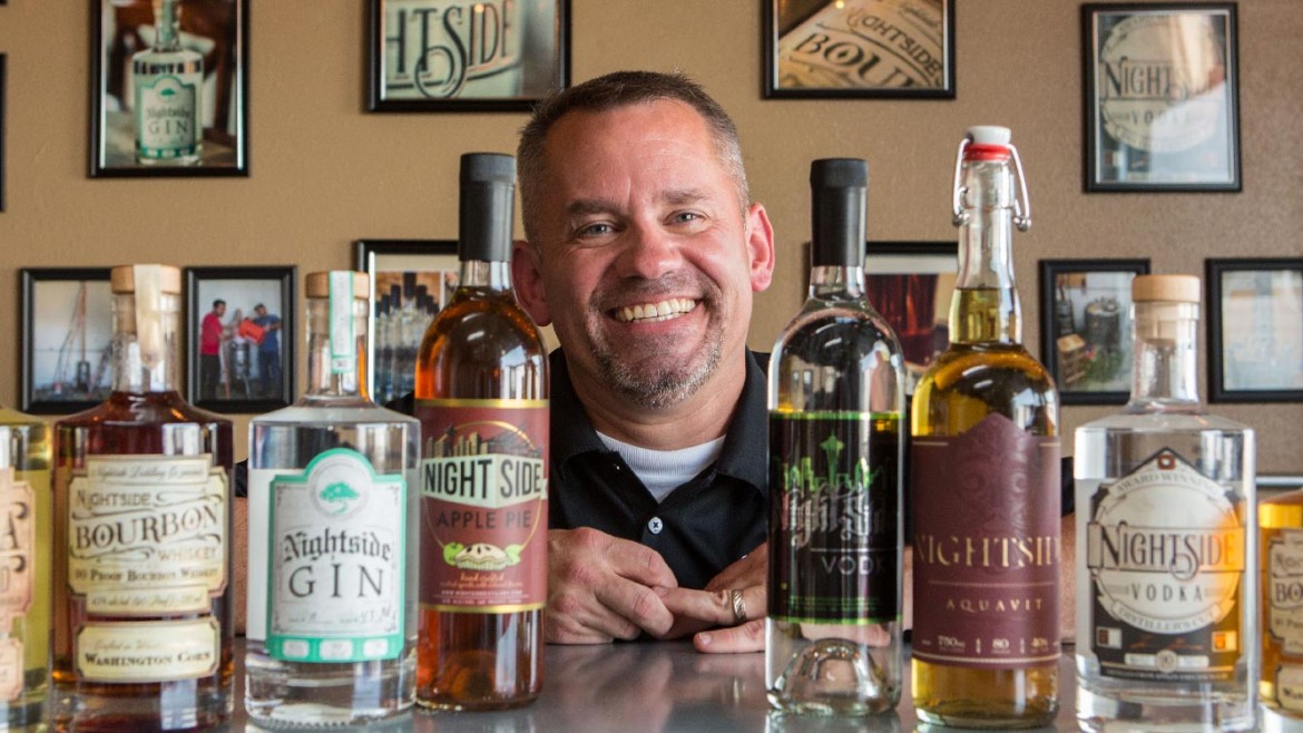 Raymond Bunk smiling with his bottles of alcohol from the distillery in front on him