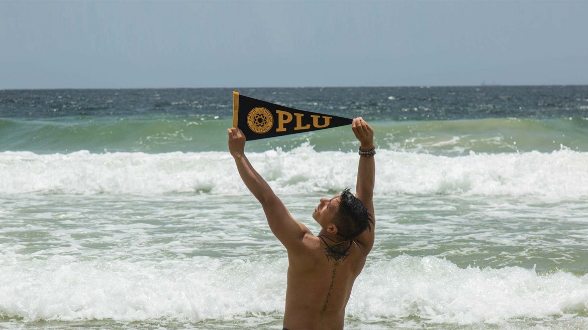 A PLU student holding a "PLU" pennant above his head on a beach