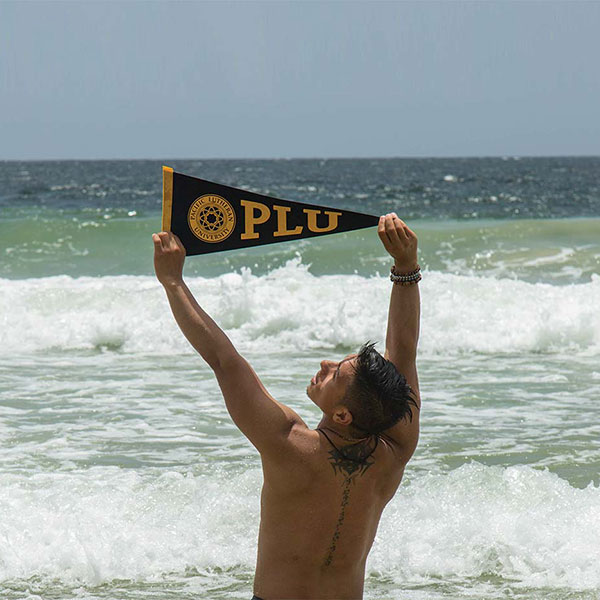 A PLU student holding a "PLU" pennant above his head on a beach