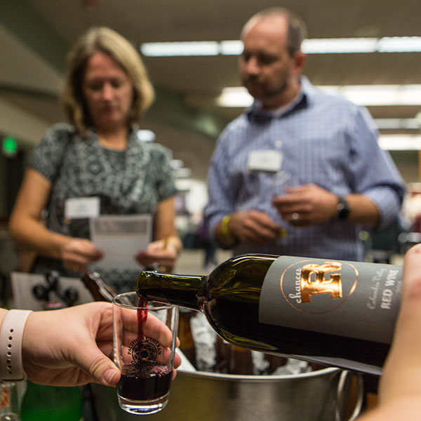 Someone pouring a glass of wine while two people in the background read a paper