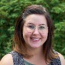 Kelsey is standing in front of a green bush wearing round glasses and smiling.