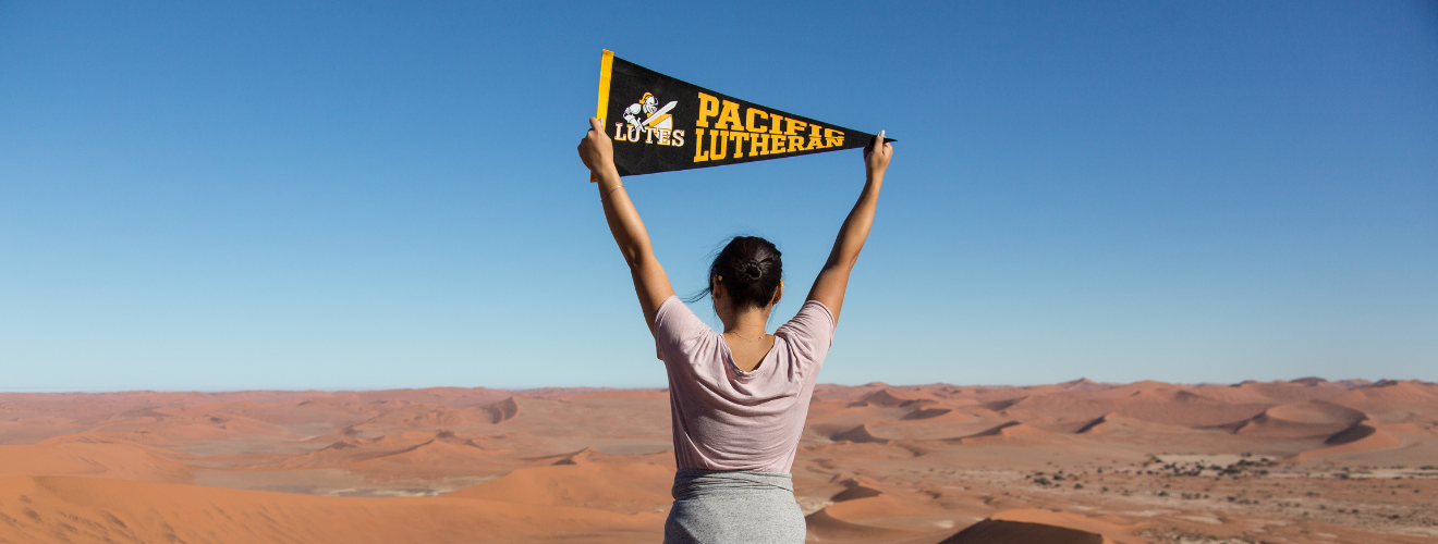 Student holds a PLU penant above her head with sandy dunes in front of her