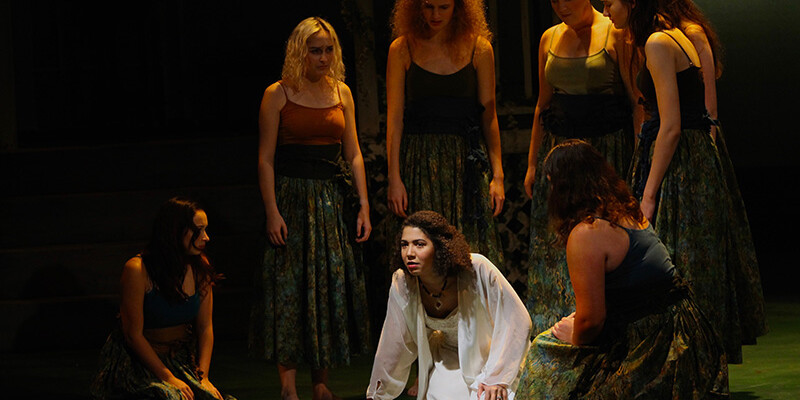 A group of female chorus members dressed in green surround Medea dressed in white.