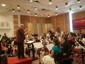 2009 China University Wind Ensemble Tour - A conductor standing in front of an orchestra, with a music stand in front and various musicians playing instruments in a concert hall with a high ceiling.
