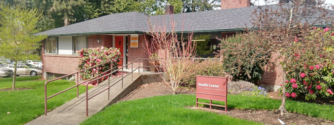 PLU Student Health Center with a red sign in the yard, surrounded by plants and trees, with a porch and a walkway leading up to it.