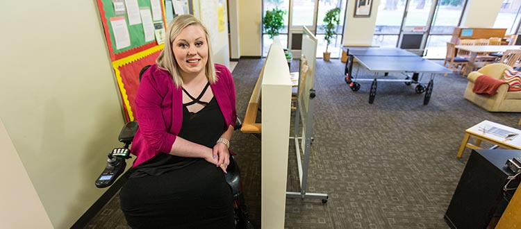 Noelle Green smiling and posing for a picture indoors, seated on a chair beside a whiteboard and artwork on the wall.