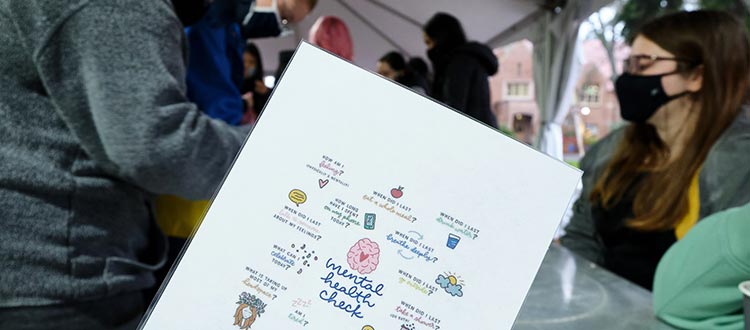 A woman standing and looking at a sign with handwritten text indicating "mental health check." She is dressed in casual clothing.